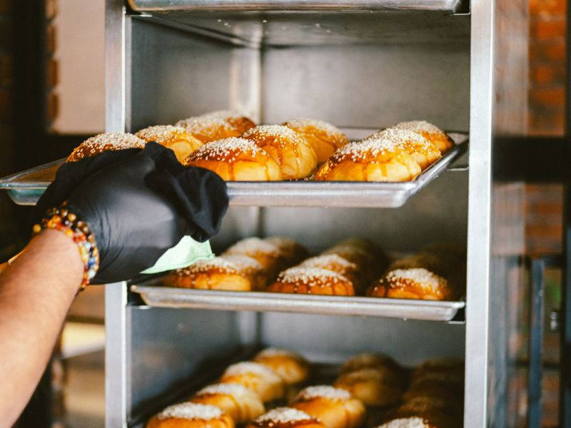 A gloved hand sliding a tray of fresh buns from a bakery rack