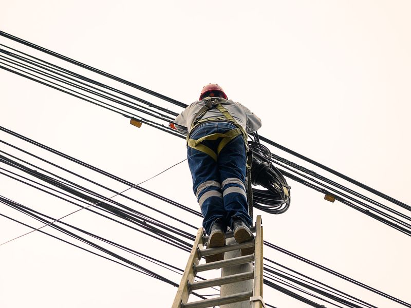 A technician on a ladder working on overhead lines against a pale sky