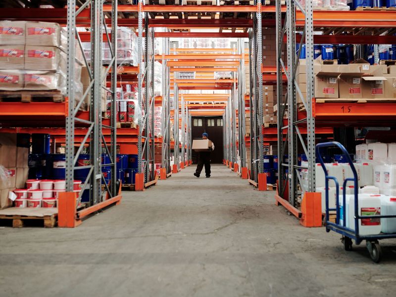 A worker carrying a box down a warehouse aisle of stocked racking