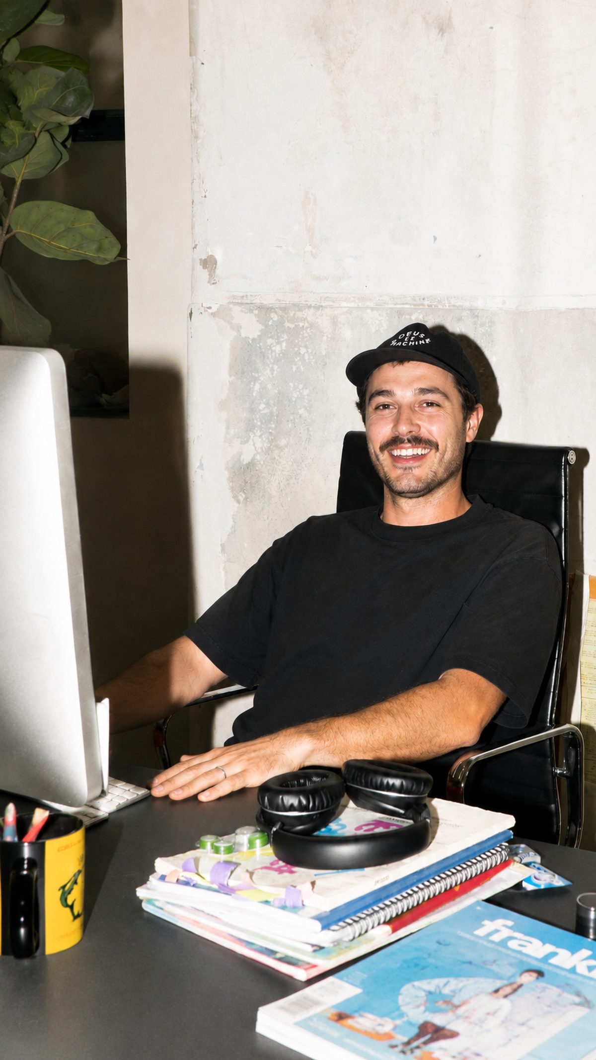 A smiling man at a creative studio desk, wearing a cap